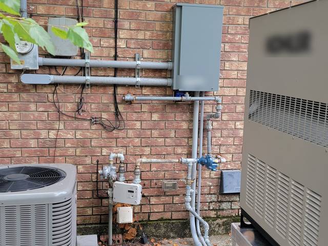 A standby generator beside an AC unit, with its transfer switch on the brick wall